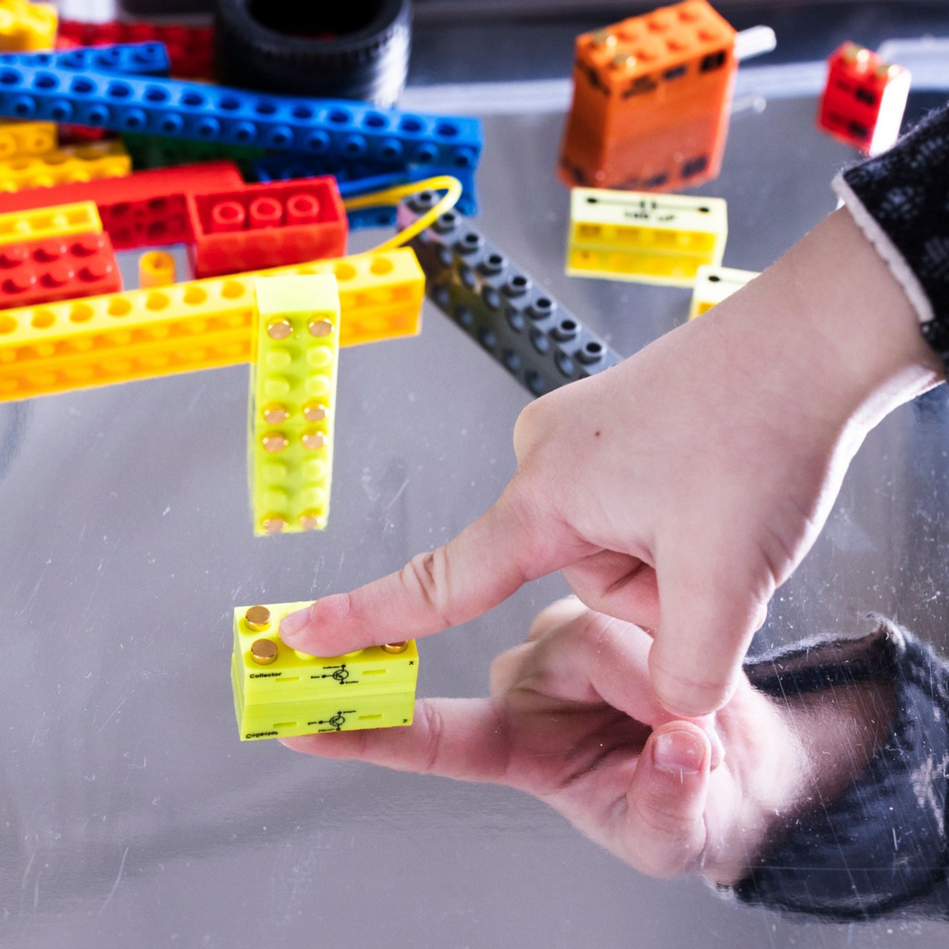 Hand touching a lime green building block with printing on the side showing it is a functional transistor with colorful LEGO pieces in the background on a reflective surface.