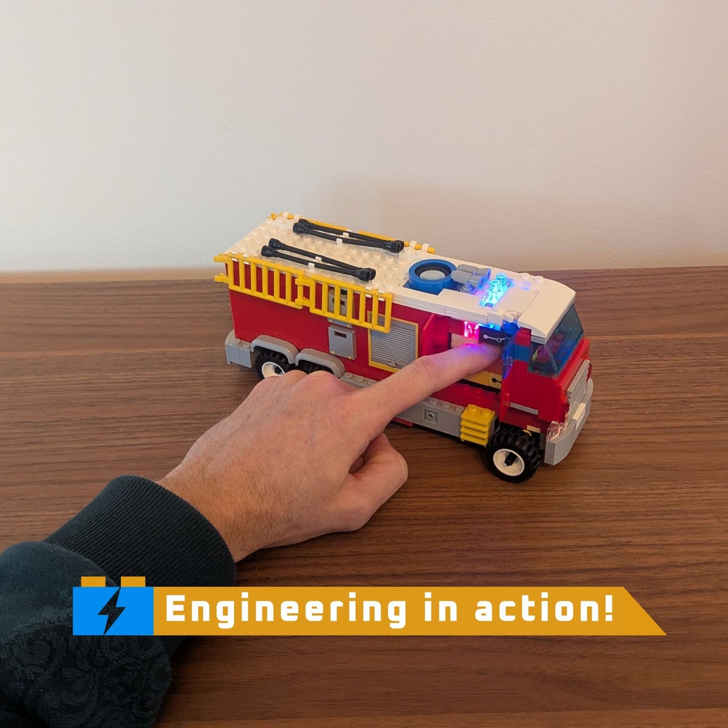 Toy fire truck with a hand pressing a button to light lights on a wooden surface, featuring 'Engineering in action!' text.