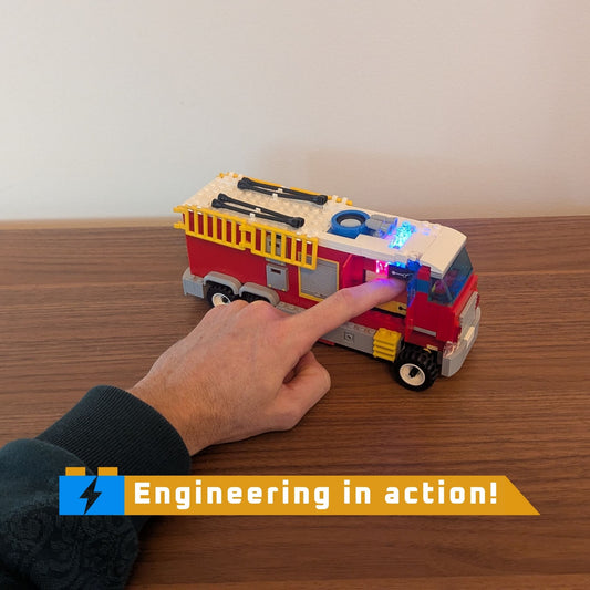 Toy fire truck with a hand pressing a button to light lights on a wooden surface, featuring 'Engineering in action!' text.