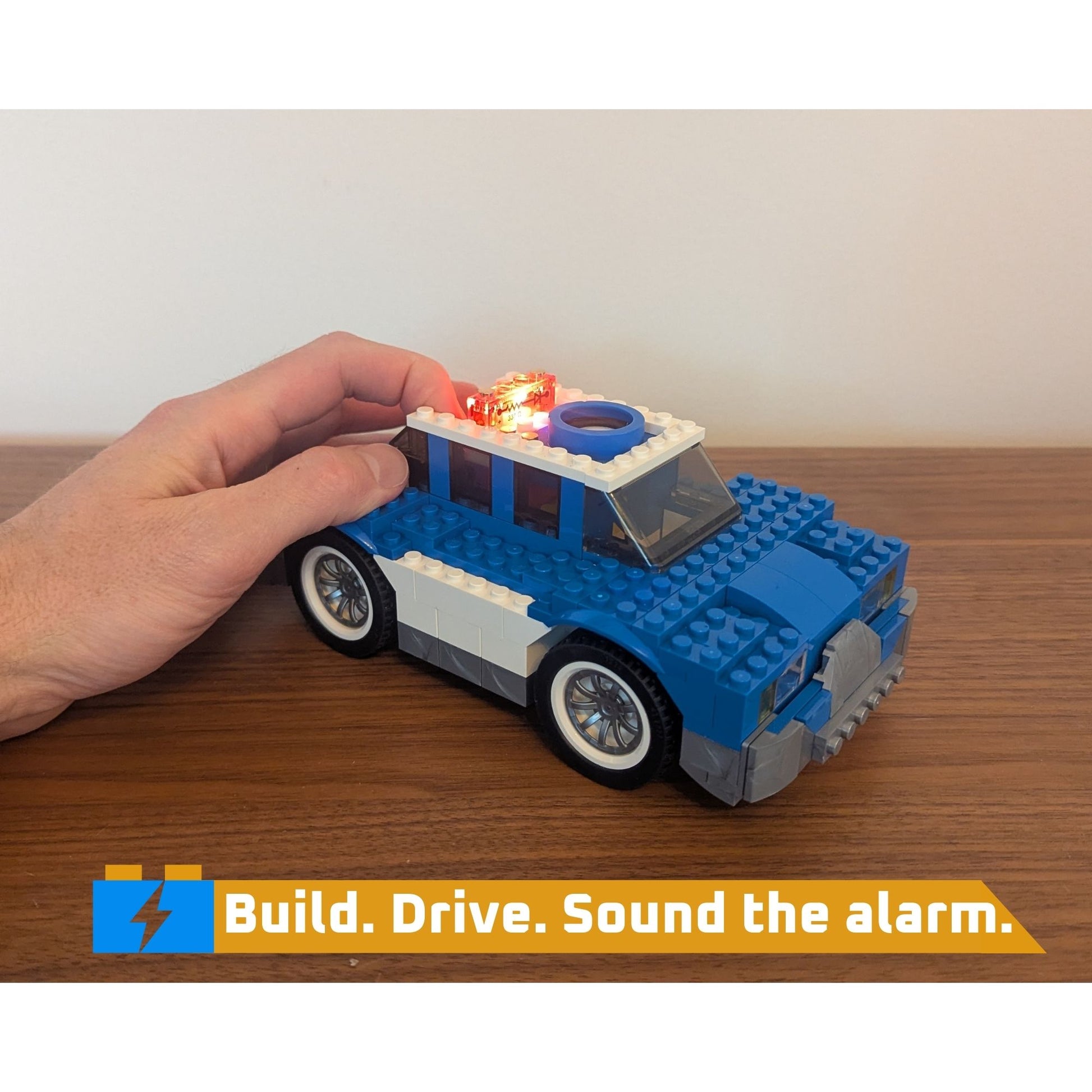 Hand holding pushing a button on a blue and gray toy patrol car with lights and speaker on a wooden surface, featuring text 'Build. Drive. Sound the alarm.'