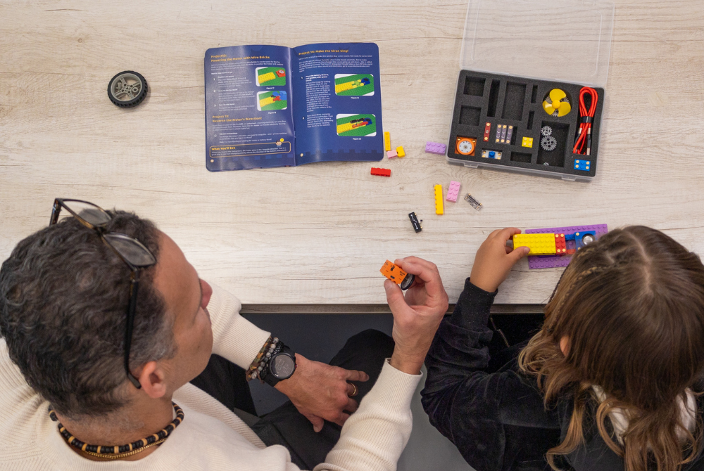 An overhead view of a man and a child assembling a small toy or educational kit on a light wood tabletop. The scene includes an open instruction booklet, loose small parts, and a clear plastic case containing the kit components.