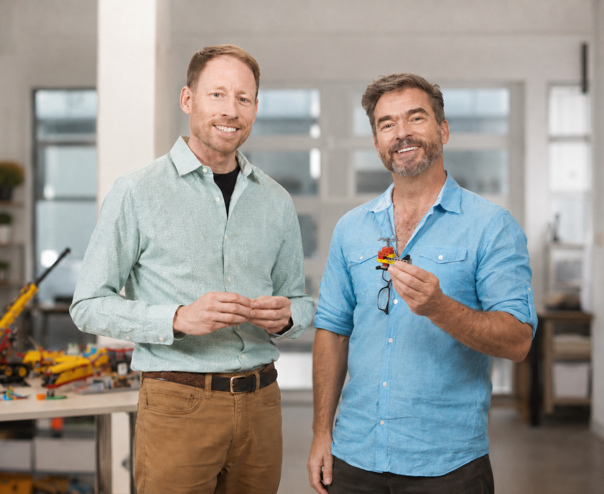 Two men standing in a bright, modern workshop or studio. The man on the right holds up a small, colorful object that appears to be a miniature toy or model made from LEGO bricks and glasses.