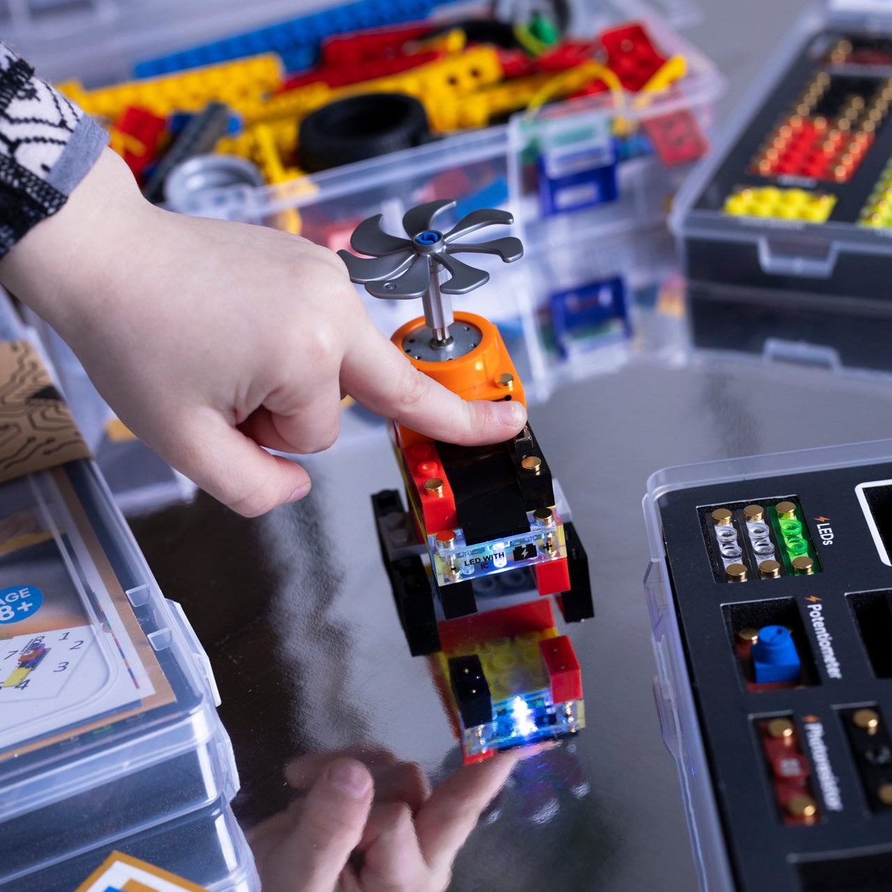 Child's hand holding a small robot model with a fan on top, surrounded by educational materials including 'SparkBlocks' packaging.