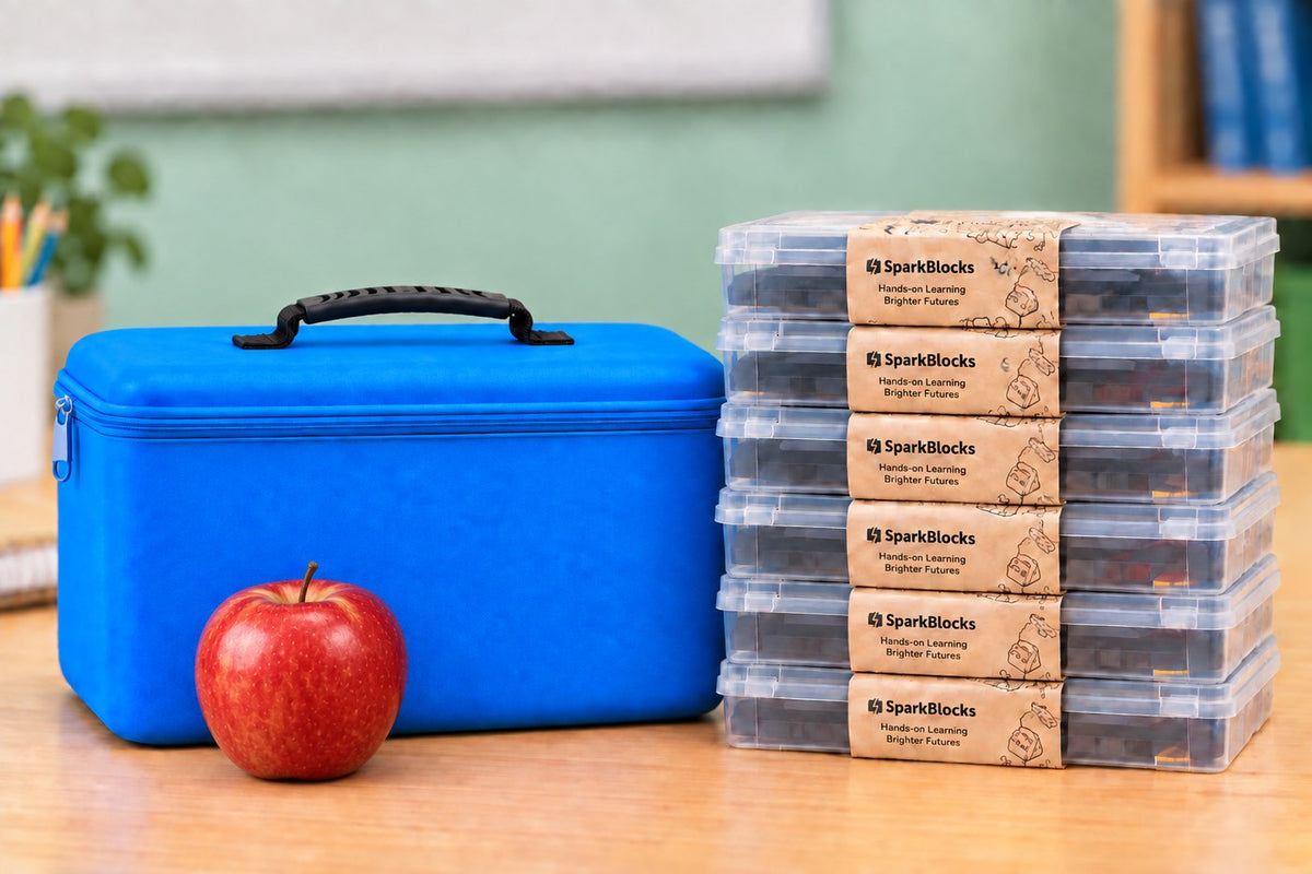 The blue SparkBlocks carrying case beside a stack of six banded kits and an apple on a classroom desk