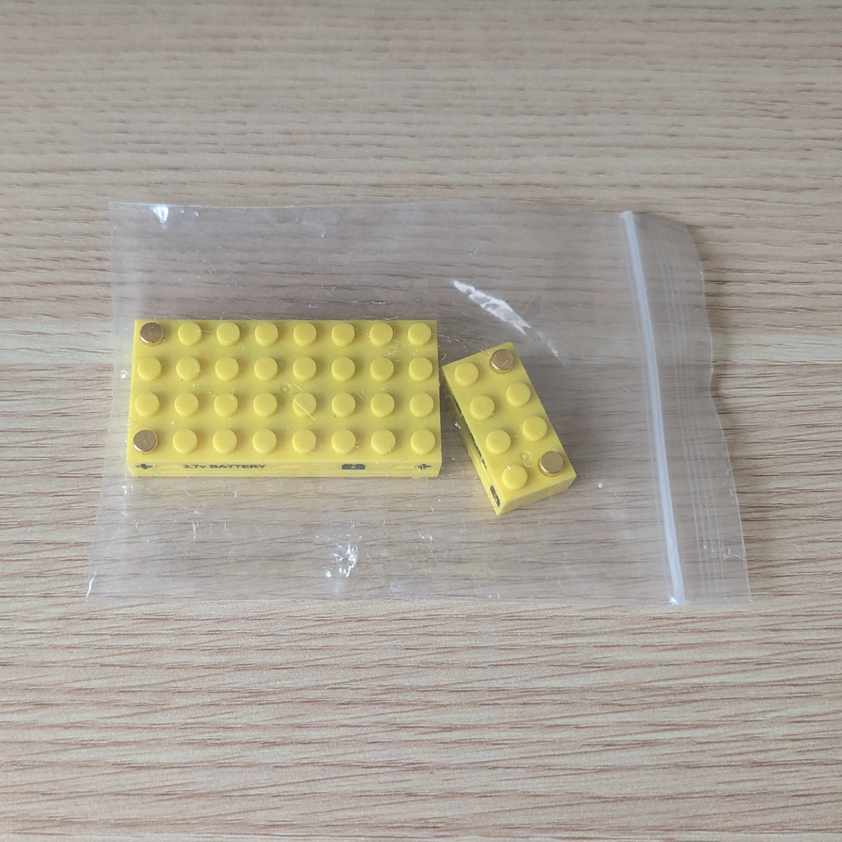 Two yellow building blocks in a clear plastic bag on a wooden surface: one rechargeable battery block and one USB-C power block
