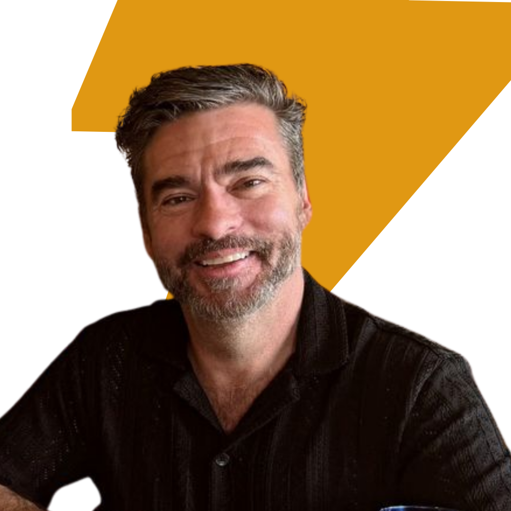 A smiling man with graying hair and a beard wearing a black textured collared shirt, set against a graphic background with a large mustard yellow shape on a white field.