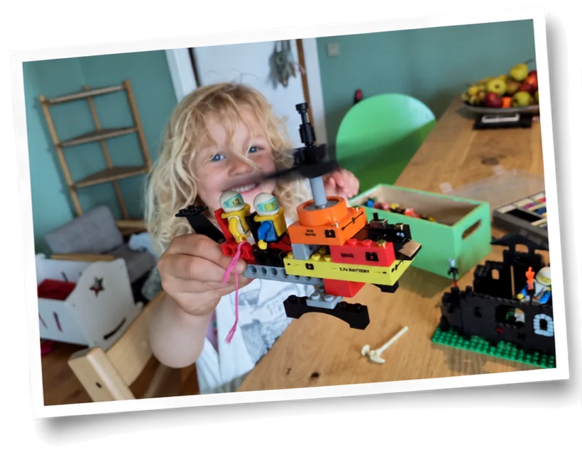 A child smiles while holding up a SparkBlocks build at a table